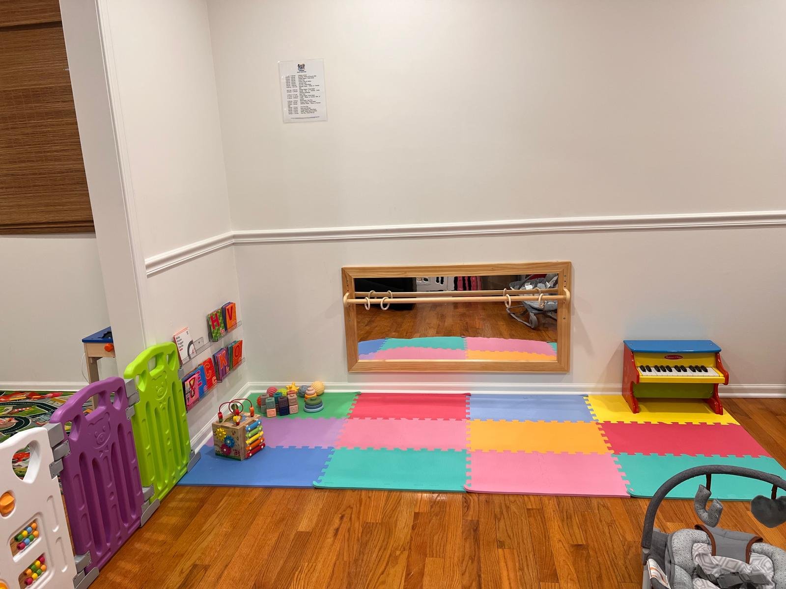 Children's play area with colorful foam mats on hardwood floor, small plastic fencing, toys including a dollhouse, a toy piano, and a activity cube, with mirror on wall and a small bar for toddlers.