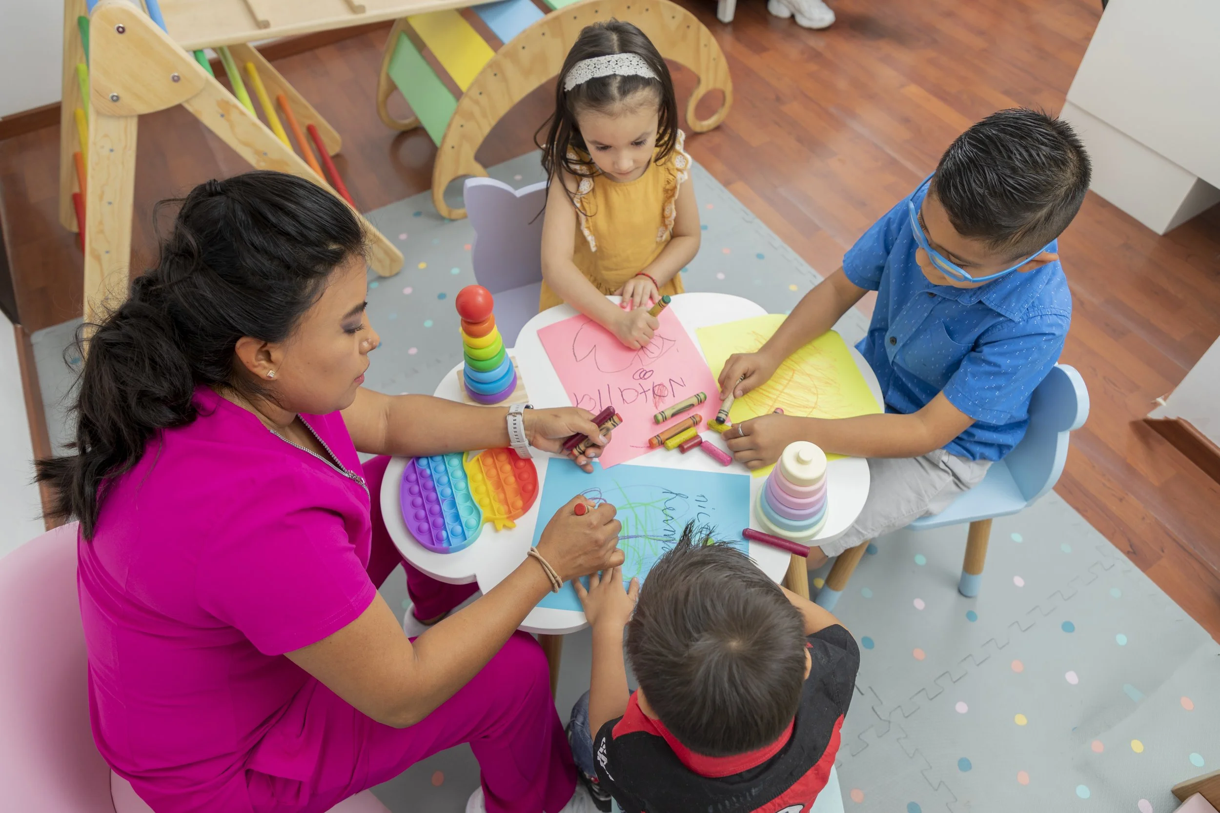 A woman and three children sitting around a small table, drawing on colorful paper with crayons in a classroom or playroom. The table has a rainbow pop-it fidget toy and a stack of cups. One child is drawing a flower on pink paper, another on blue paper, with the woman and other children engaged.