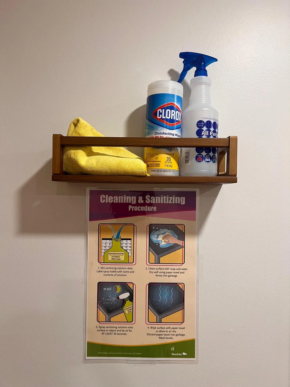 A wall-mounted shelf with cleaning supplies including Clorox disinfectant wipes, a spray bottle, a yellow cleaning cloth, and a container of wipes. Below the shelf, a poster displays cleaning and sanitizing procedures with illustrated steps and text.