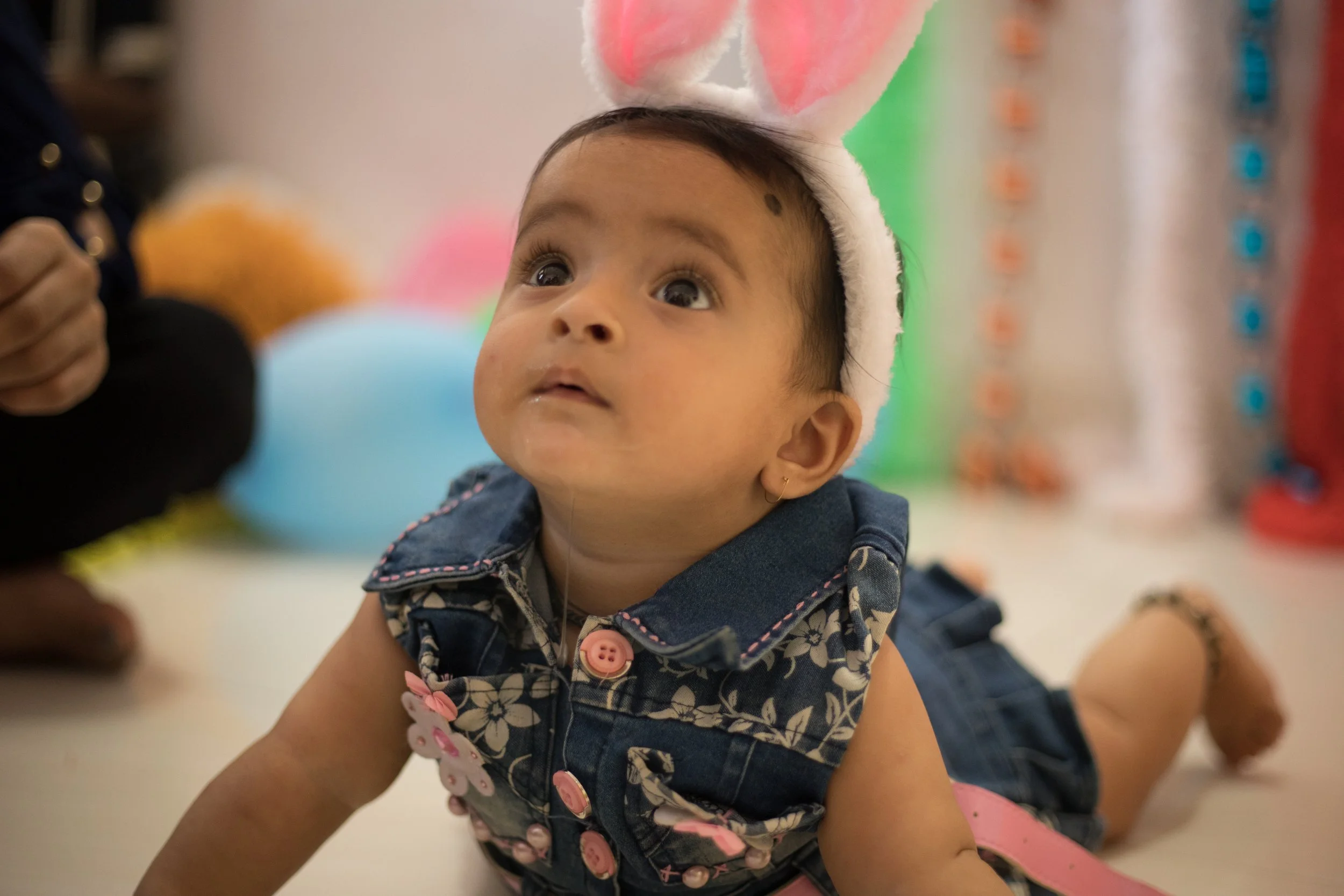 A young girl wearing bunny ears and a denim dress, lying on her stomach on the floor, looking upward.