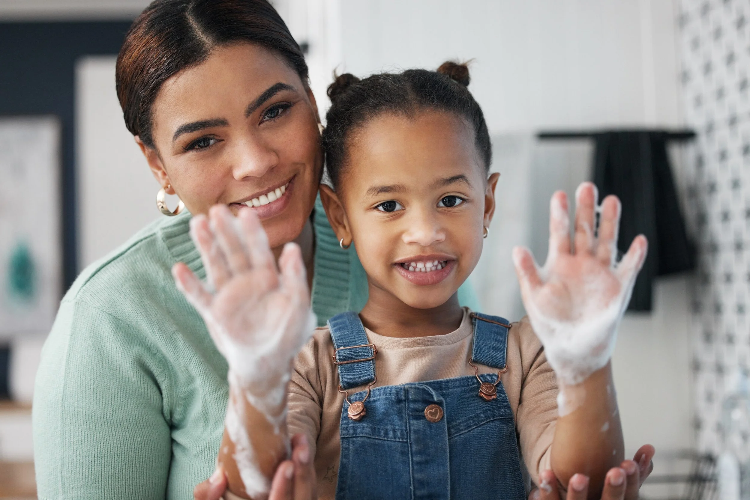 A woman and a young girl smiling and showing their soapy hands in a kitchen.
