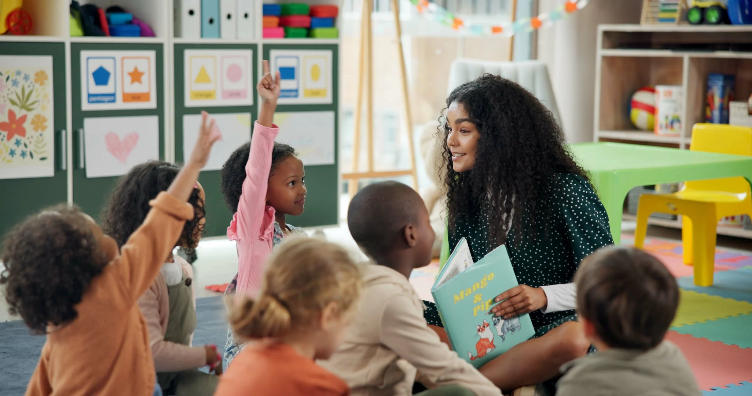 A woman reading a children's book titled 'Mango & Pi' to a group of children in a classroom. Some children are raising their hands, sitting on colorful mats.