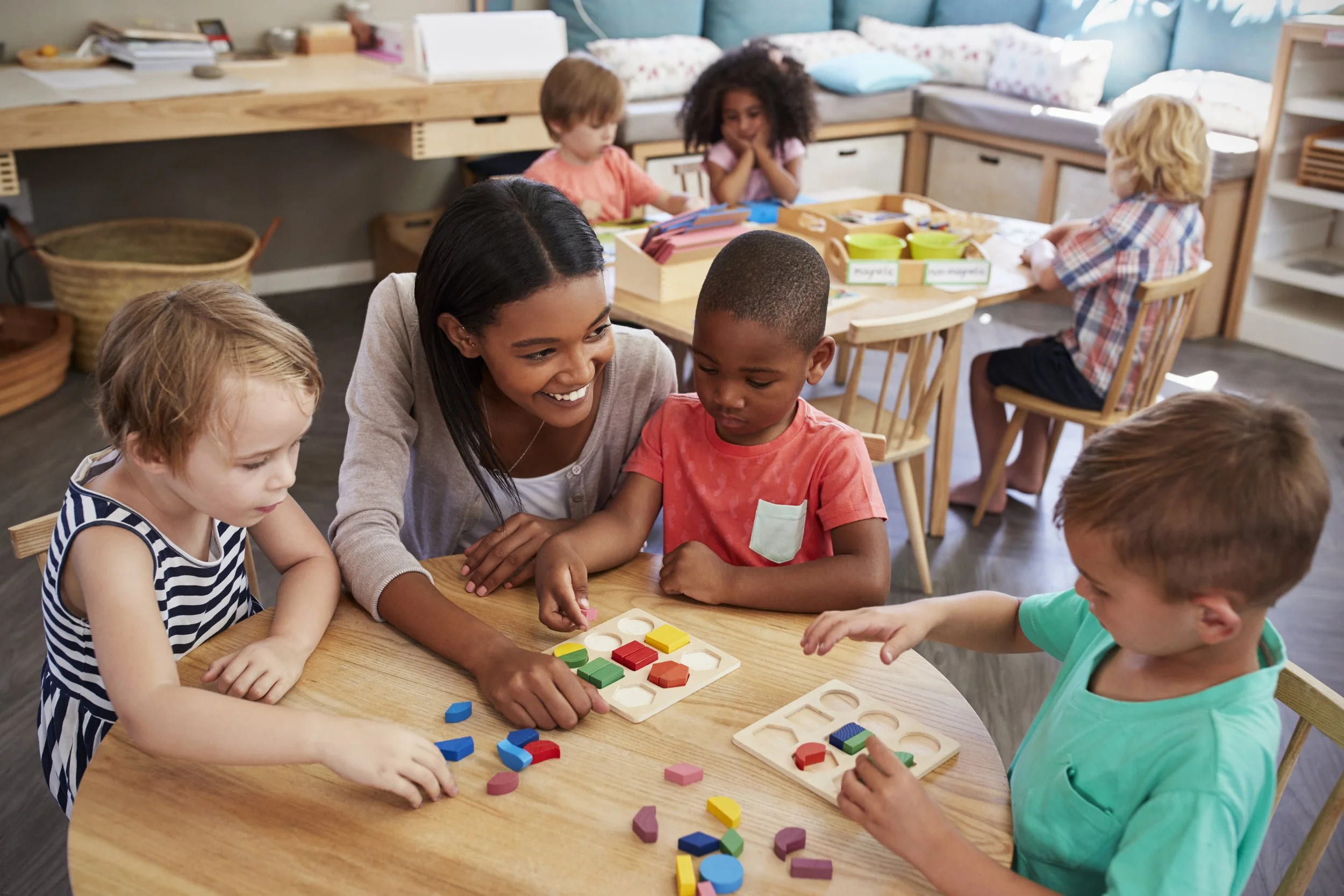 A diverse group of young children and a teacher playing with colorful geometric puzzle pieces around a wooden table in a classroom.