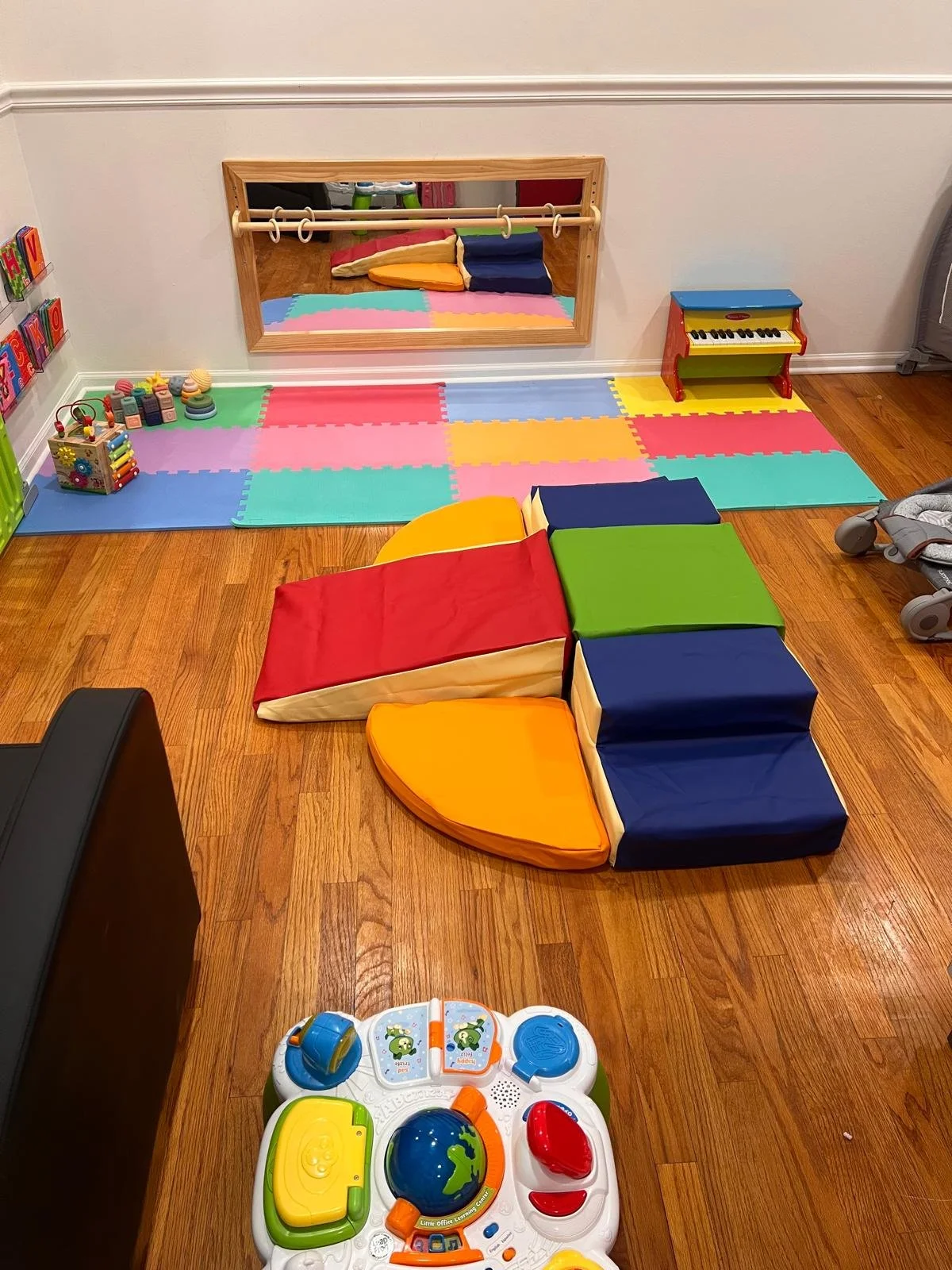 A colorful indoor play area for children with foam mats, soft blocks, a mirror, toys, and a small piano, all on a wooden floor.