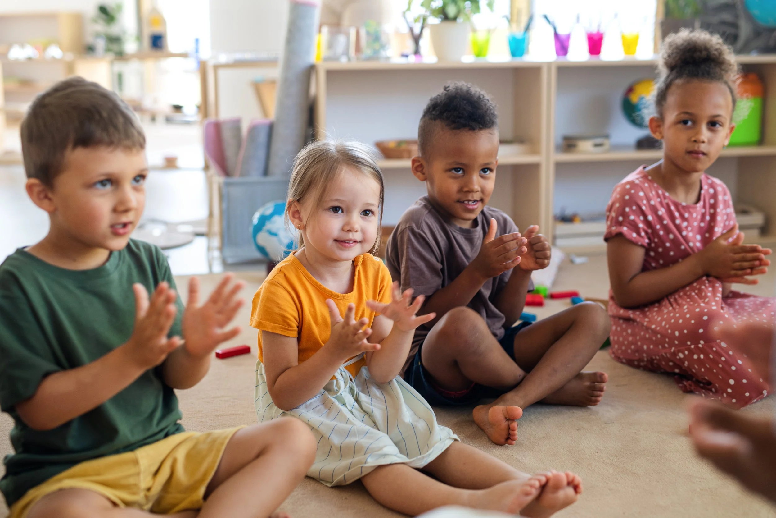 Four children sitting on the floor, clapping and smiling, in a classroom or playroom with shelves, toys, and colorful cups in the background.