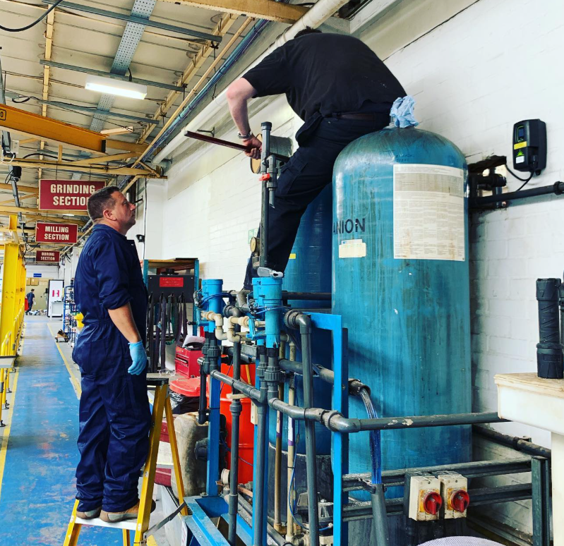 Two workers in a factory, one standing on a ladder and the other perched on a large blue tank, inspecting industrial equipment.