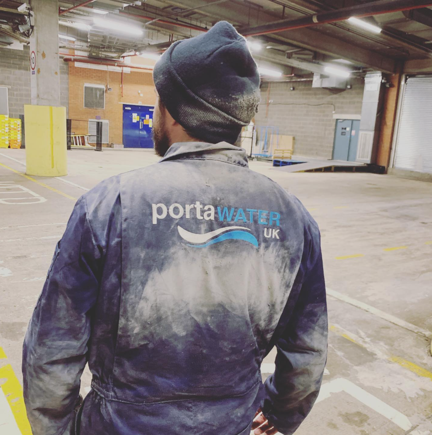 A worker in a gray jumpsuit with 'PortaWater UK' logo on the back, standing in an industrial parking garage. The worker is wearing a beanie and is facing away from the camera.
