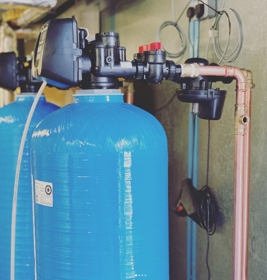 Blue filtration tank with black control valve and copper piping in a utility room.
