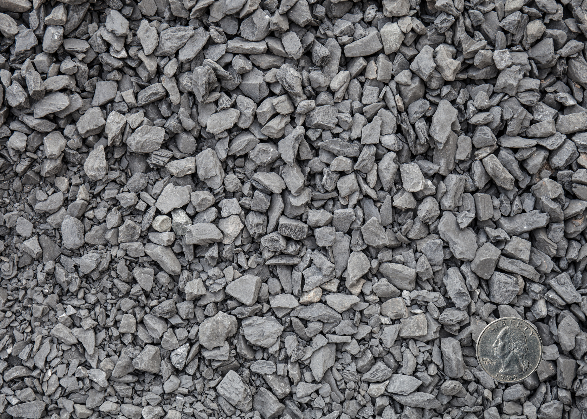 Close-up of a pile of small gray gravel stones with a quarter dollar coin for scale in the bottom right corner.
