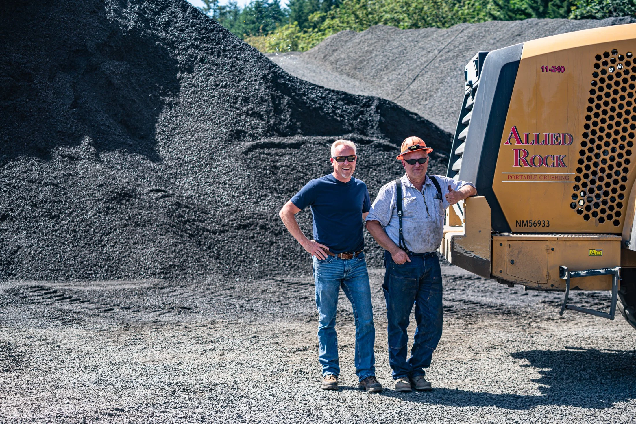 Two men standing in front of a large pile of crushed rock or gravel, next to construction equipment labeled 'Allied Rock Portable Crushing'.