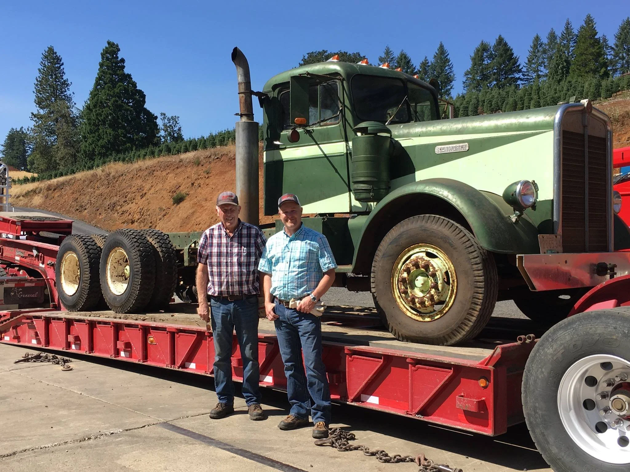 Two men standing in front of a vintage green and cream semi-truck on a red flatbed trailer, outdoors with trees and a hillside in the background.