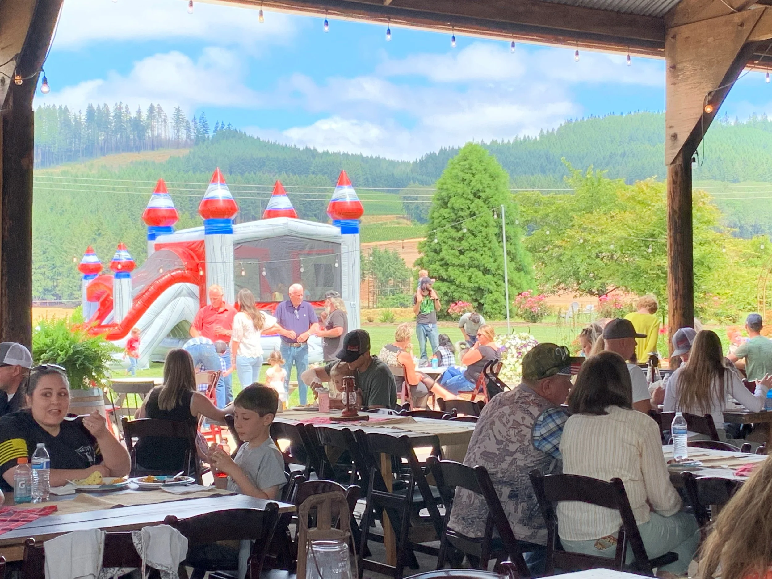 People dining at an outdoor restaurant with a view of a children's bounce house and green hills in the background.