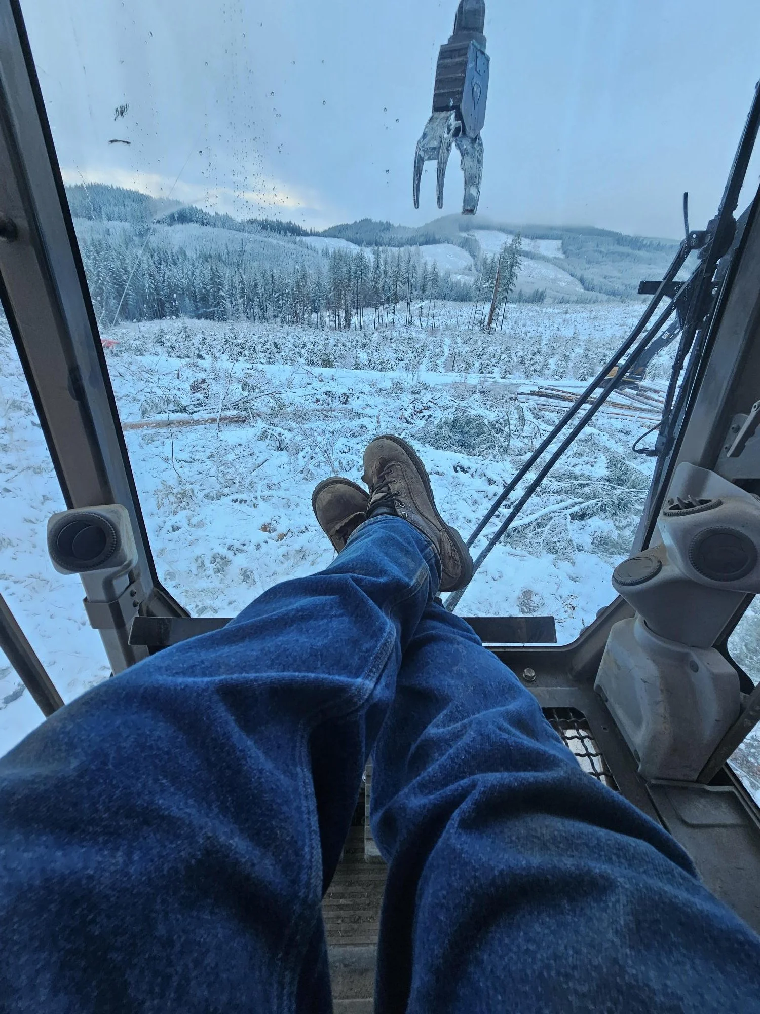 Interior view from inside a snowcat, showing person's legs and feet wearing boots, with a snow-covered landscape and forest in the background, and a large mechanical claw hanging from the ceiling.