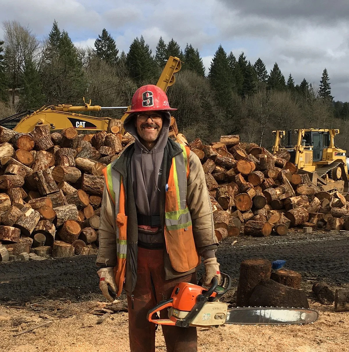 A man with glasses and a beard smiling, wearing a red hard hat with an 'S' on it, an orange safety vest, and outdoor work clothing, holding a chainsaw in front of a large pile of chopped logs and construction equipment in a forested area.