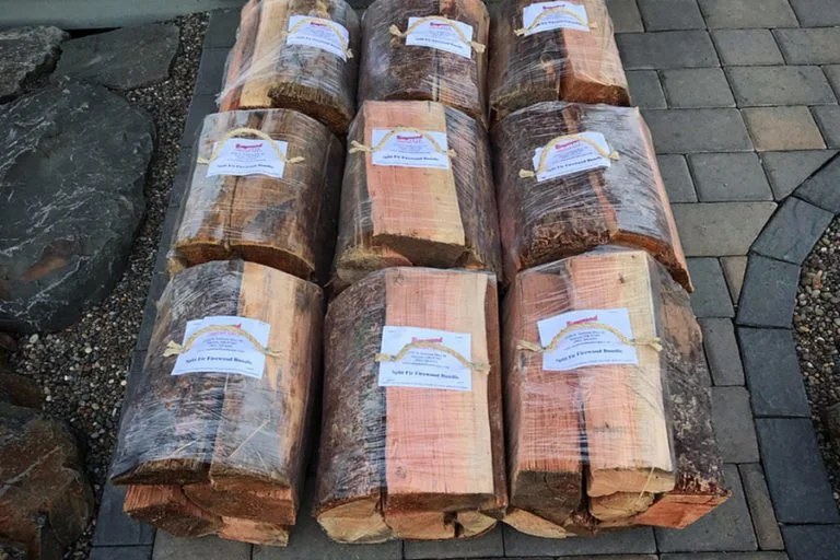 Ten bundles of firewood wrapped in plastic are arranged on a paved surface outdoors, with some rocks nearby.