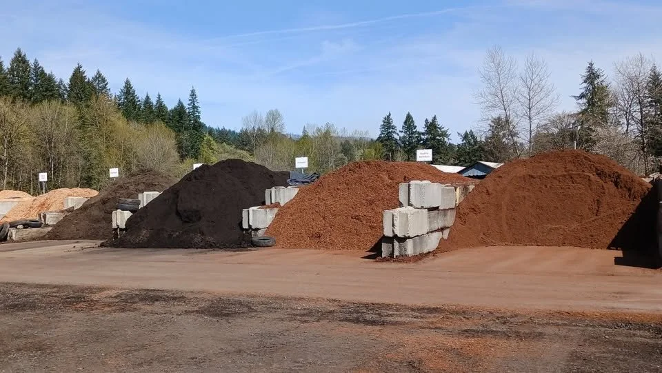 Multiple piles of different colored gravel and soil with concrete blocks in an outdoor yard, trees and a blue sky in the background.