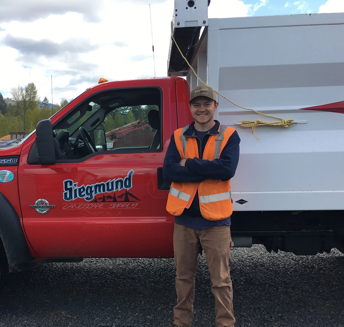 A smiling man in an orange safety vest and cap stands with arms crossed in front of a red truck with the logo 'Siegbund Landscape Supply' and a white service body. The background shows cloudy sky and trees.