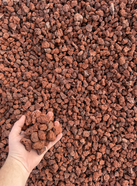 Close-up of a hand holding small, reddish-brown gravel stones, with a large pile of similar gravel in the background.