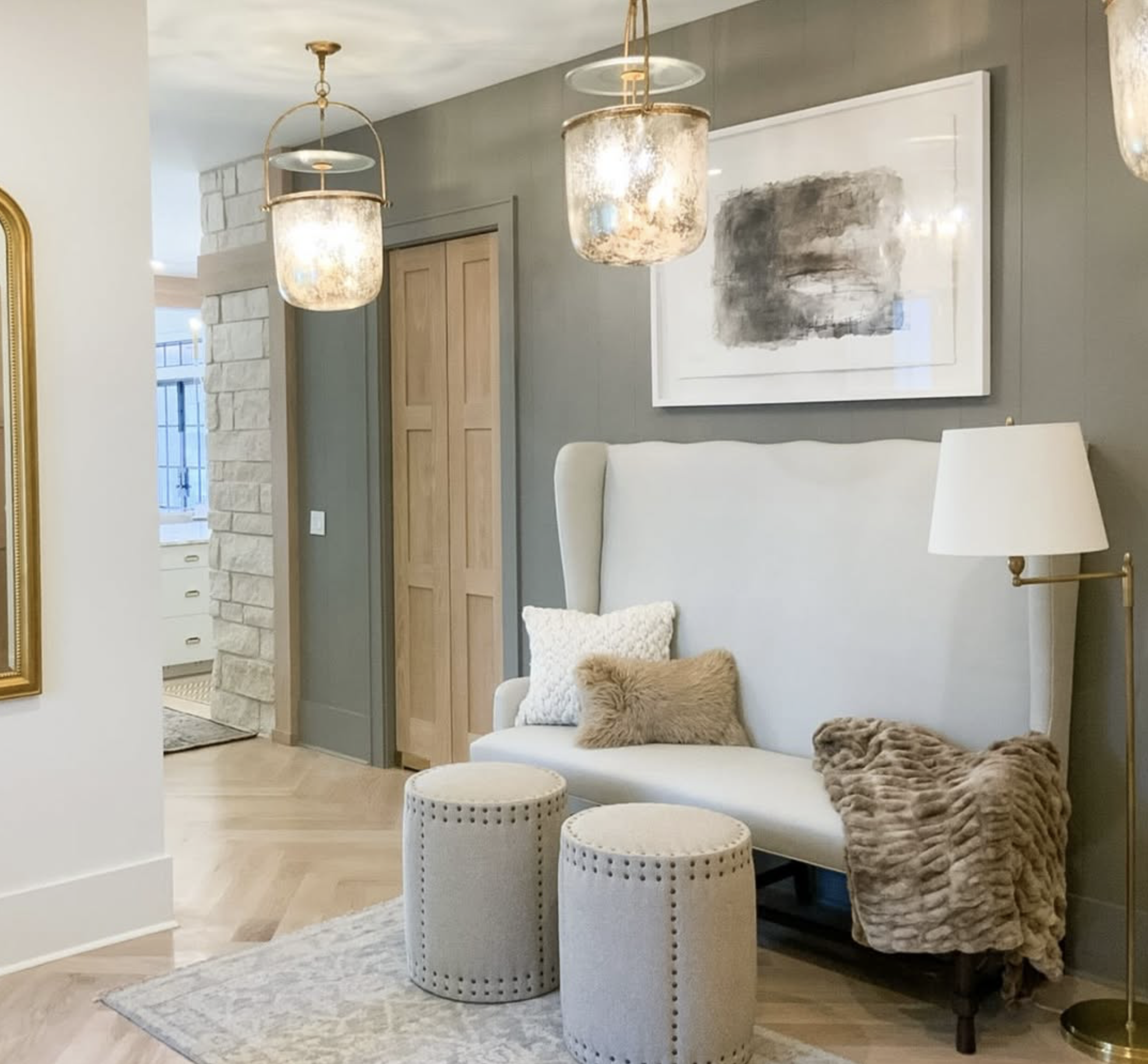 A stylish living room corner with two hanging pendant lights, a large abstract black-and-white painting, a white upholstered high-back sofa with plush pillows, a standing lamp, two beige round ottomans, and a soft faux fur blanket.