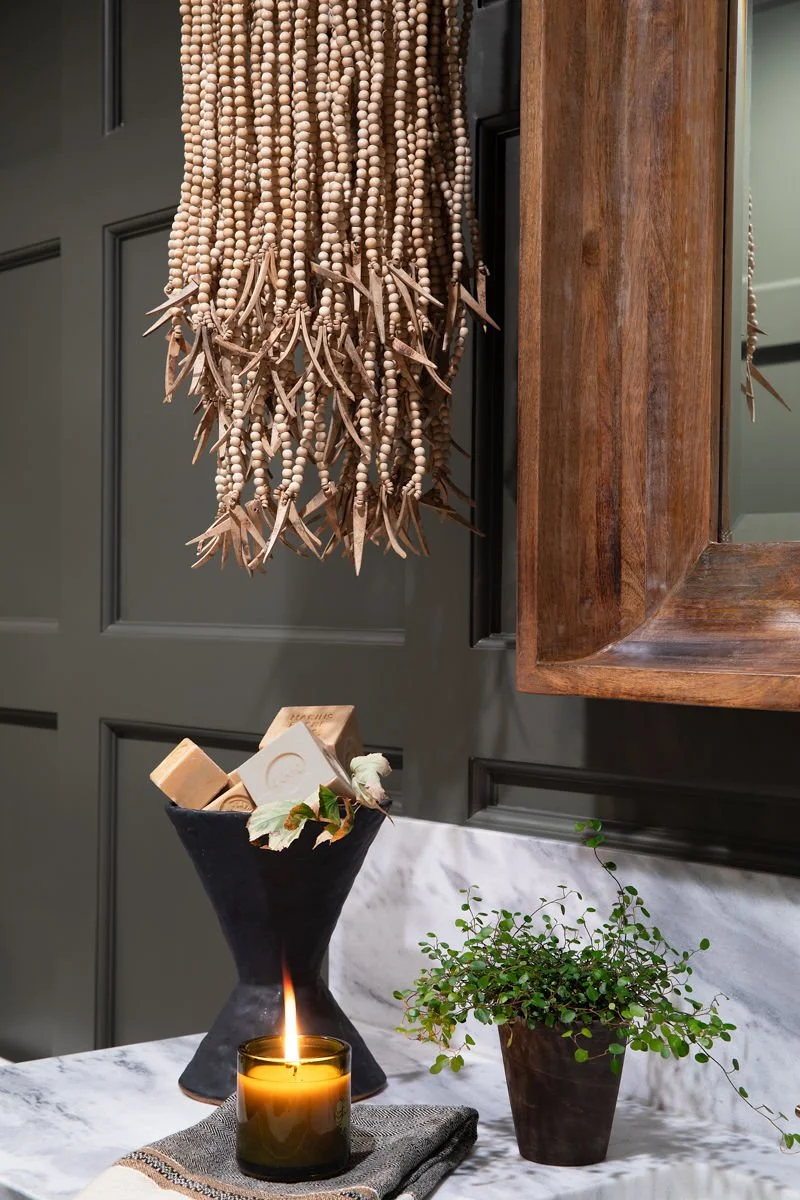 Decorative interior scene with a black candle holder with candles, a potted plant with small round leaves, a black and beige vase filled with wooden blocks and dried flowers, a hanging beaded curtain with wooden beads and star-shaped wooden ornaments, a dark green wall with paneling, and a wooden-framed mirror.