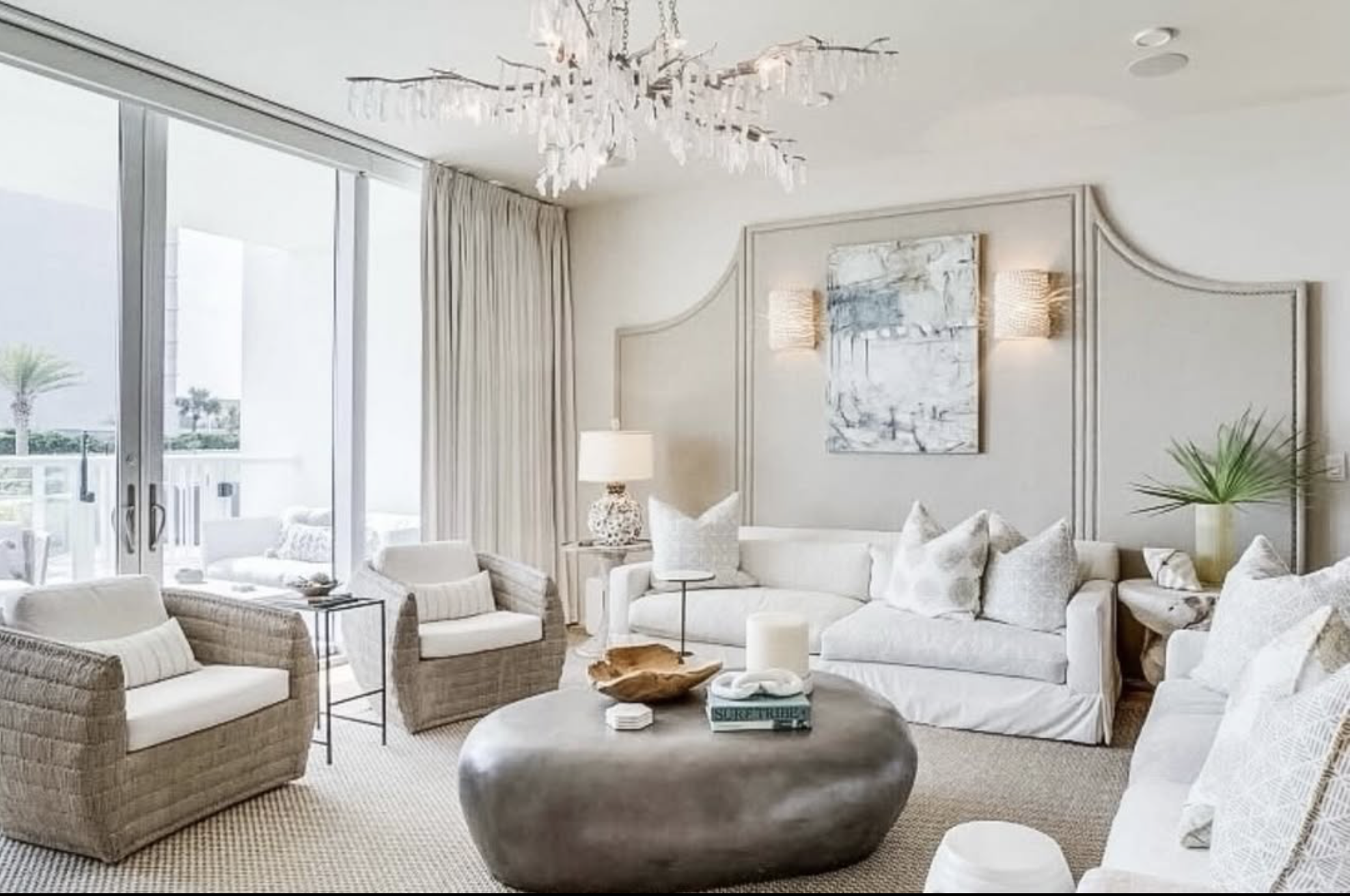 Bright living room with white sofas, beige armchairs, and a round coffee table in front of large windows with white curtains. Decorative pillows and art hang on the wall, with a chandelier overhead.