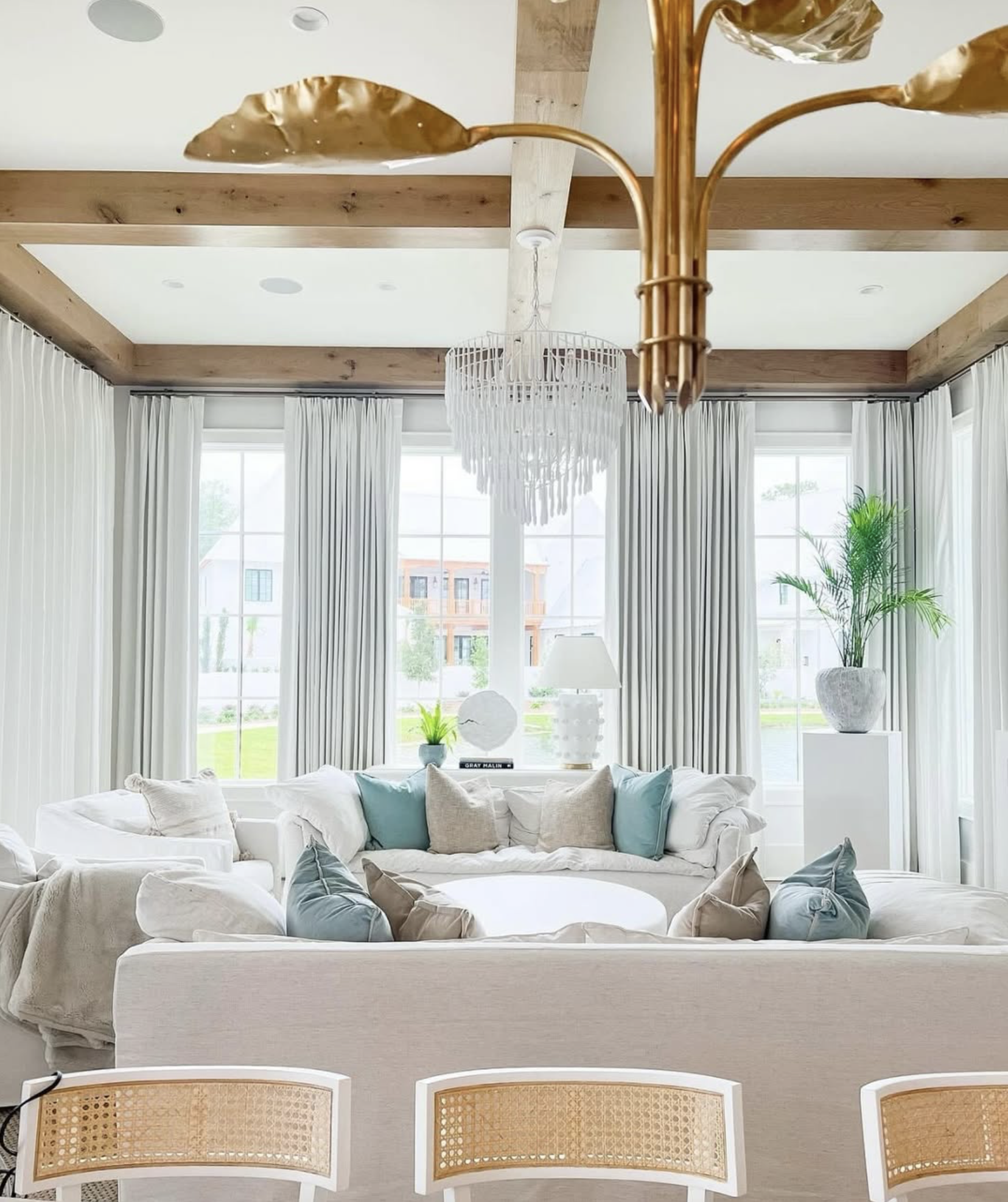 Bright living room with large windows, white curtains, white sofa, multiple decorative pillows, a potted plant, a table lamp, and a chandelier hanging from a wood-beamed ceiling.