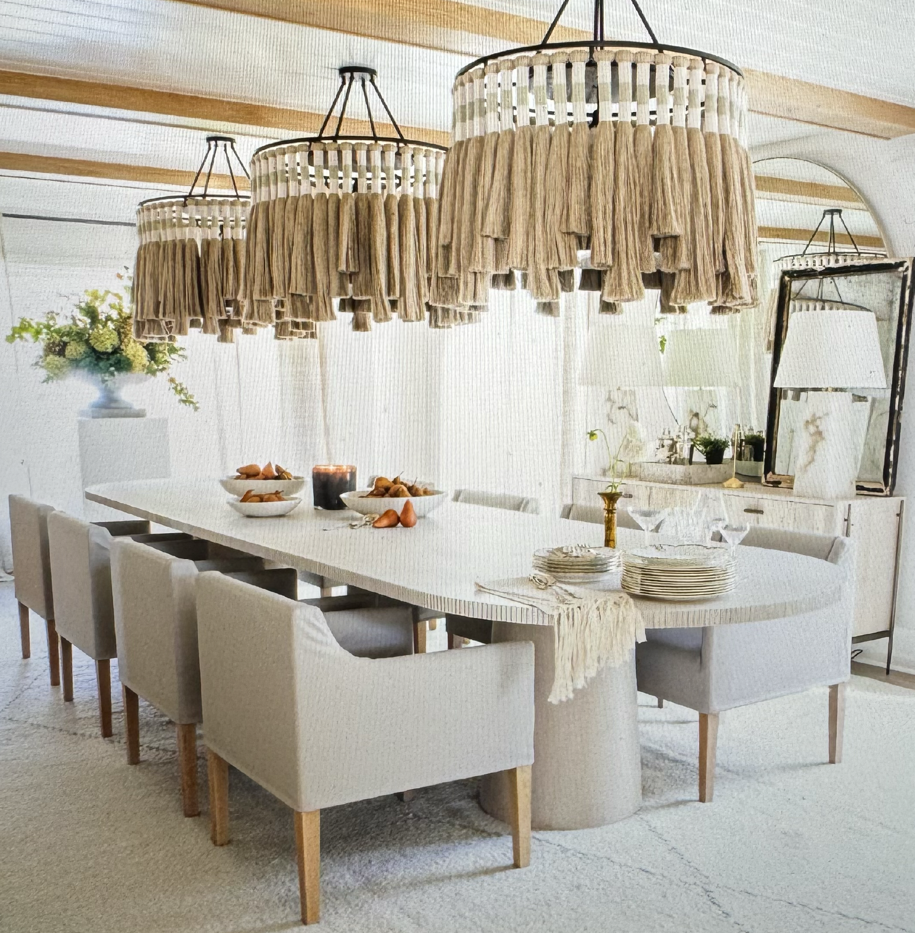 A bright dining room with a white oval table surrounded by beige chairs, large hanging wicker light fixtures, and decorative items like bowls, plates, and vases with an elegant beachy feel.
