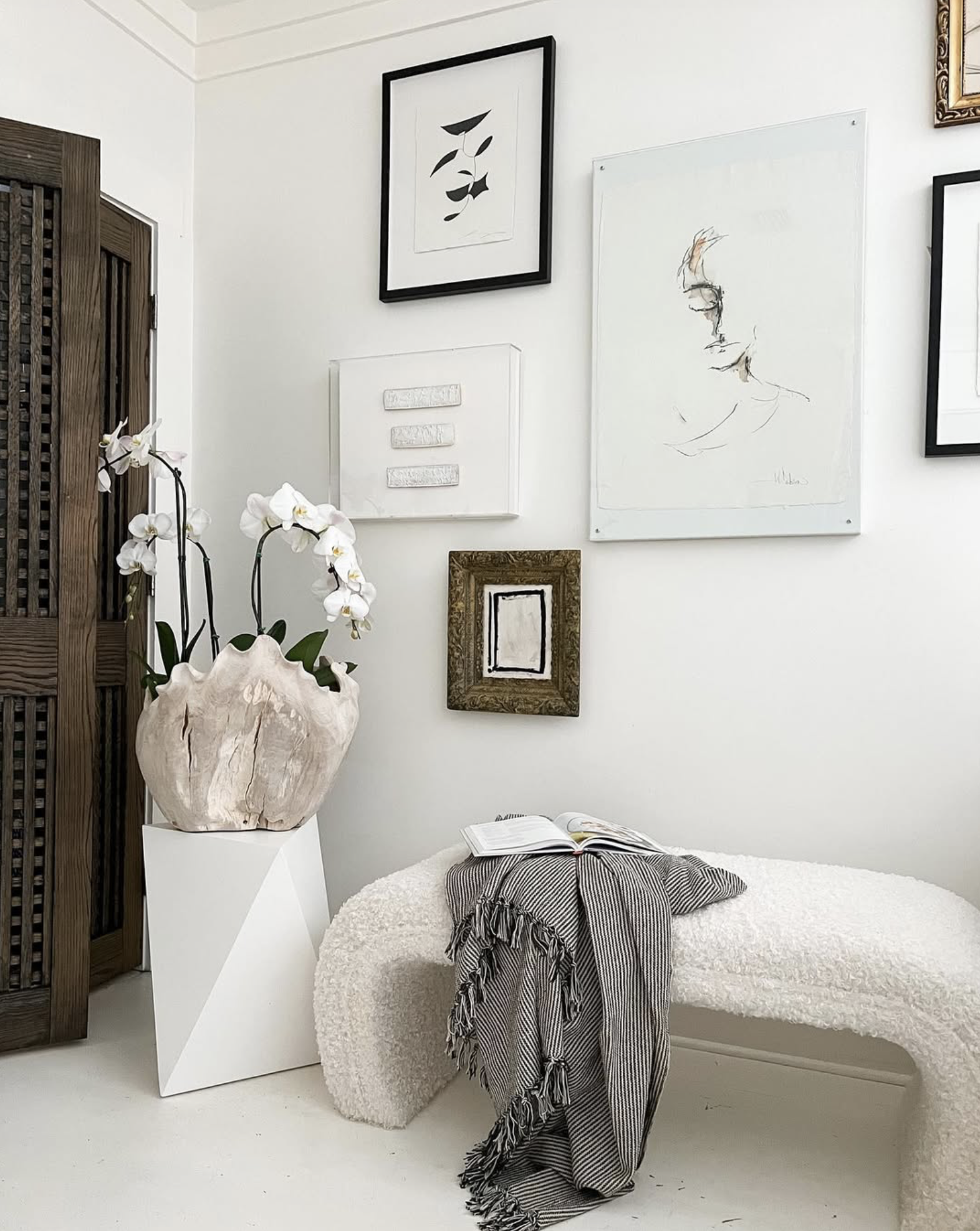 A cozy corner with a white textured bench, a striped blanket, and an open book on top. To the left, a large orchid plant in a decorative wooden vase on a white pedestal. The wall above features a gallery of abstract framed artwork and sketches in black and white.