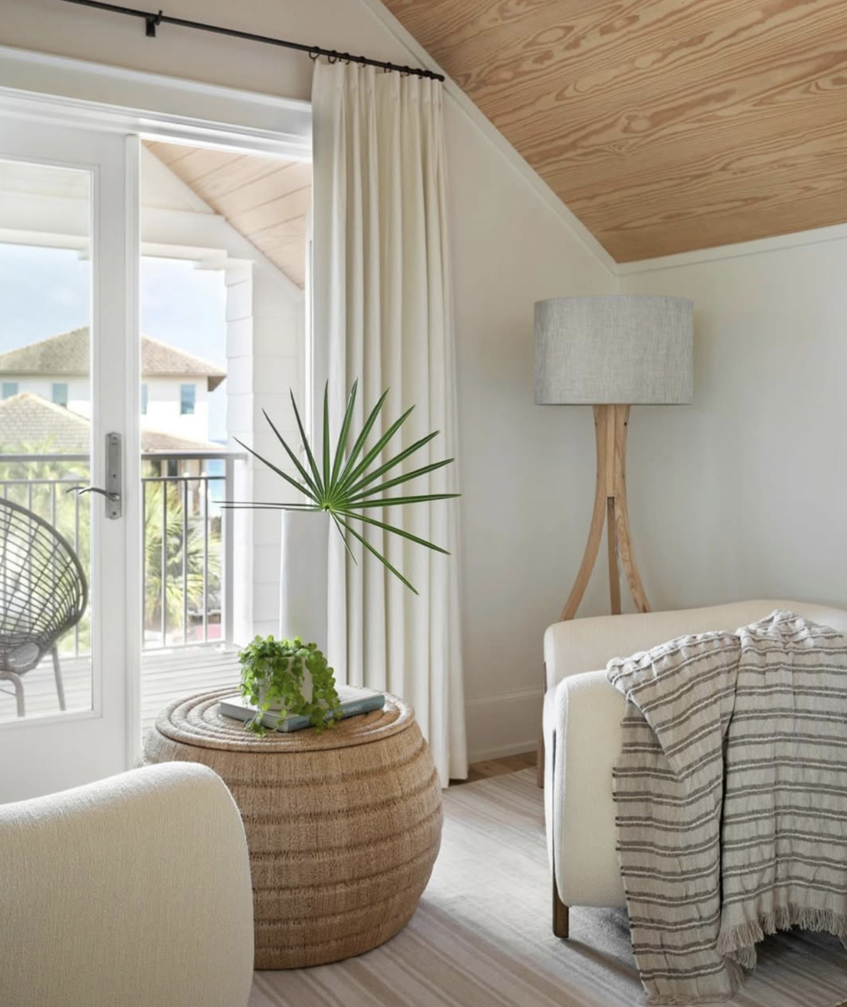 A bright living room with a beige sofa, striped throw blanket, tall wooden floor lamp, a round wicker side table with books and a potted plant, white curtains, and sliding glass doors leading to a balcony with outdoor chairs, overlooking neighboring houses.