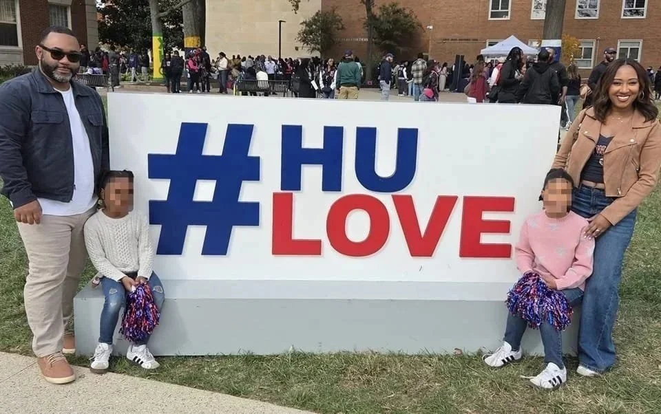 Two adults and two children posing outdoors next to a large white sign that reads '#HU LOVE' with a crowd of people and trees in the background.