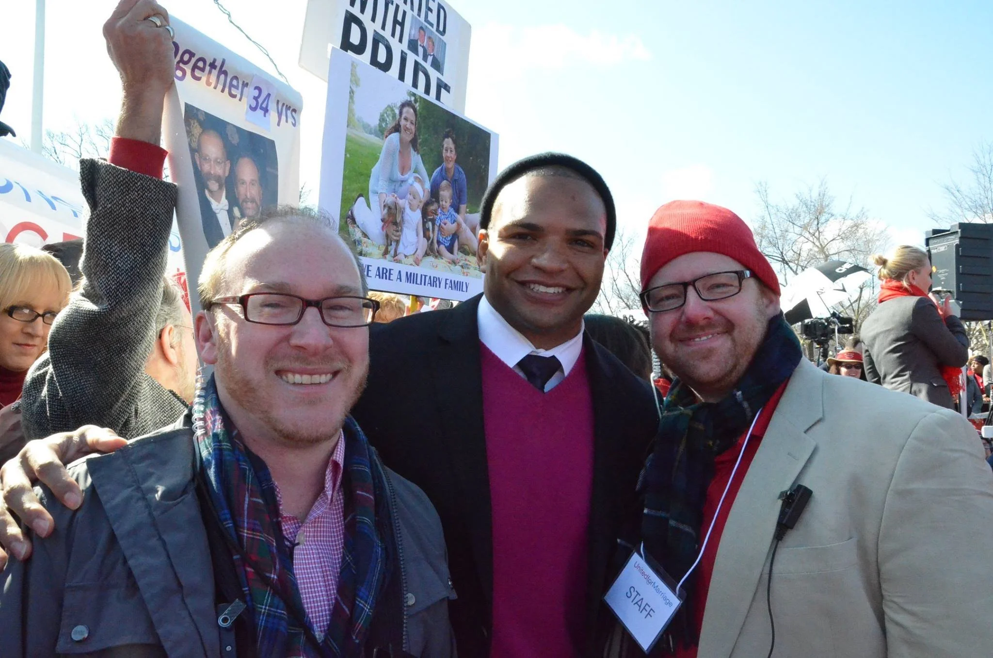Three men smiling at a gathering or protest, with signs and a crowd in the background. One man has his arm around another, and all appear happy. The signs include a photo of a woman and a message about being a military family.