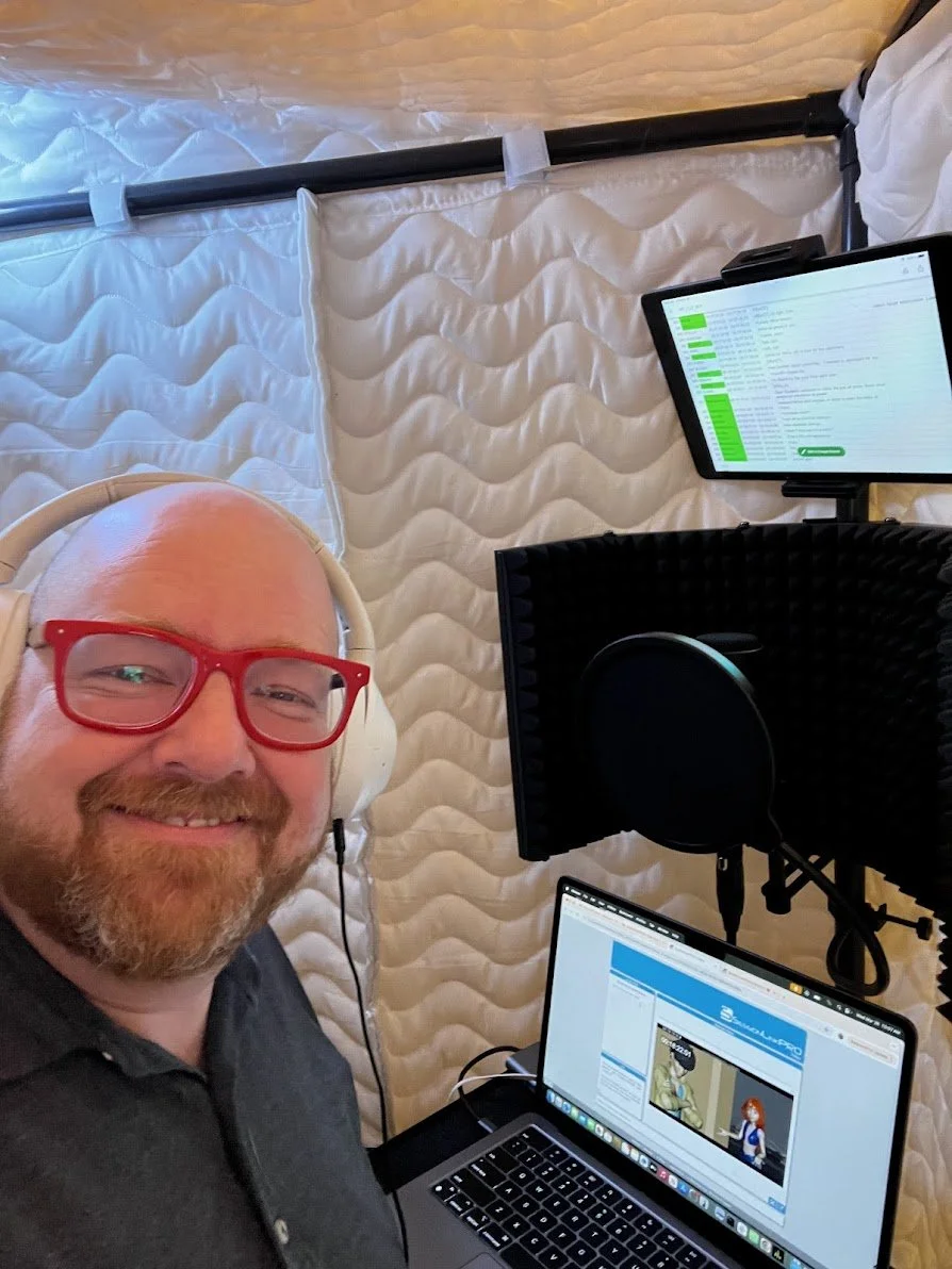 A man with a beard wearing red glasses and a black shirt taking a selfie in a recording studio. He is smiling and wearing white headphones. Behind him are soundproof walls, a laptop displaying a webpage with cartoon characters, a music monitor, a microphone with a pop filter, and a tablet showing an email inbox.
