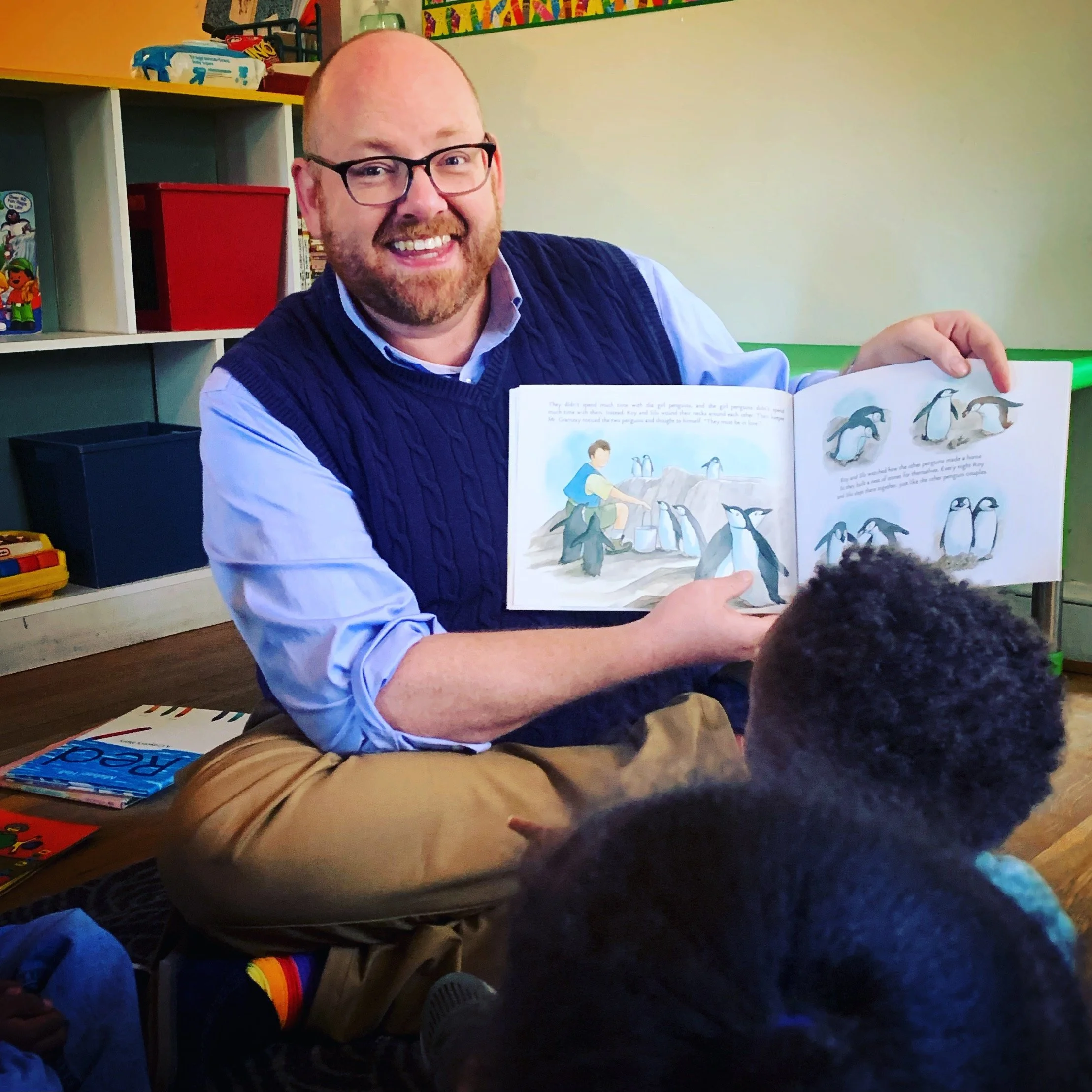 Jamie showing a children's book about penguins to a group of children in a classroom.