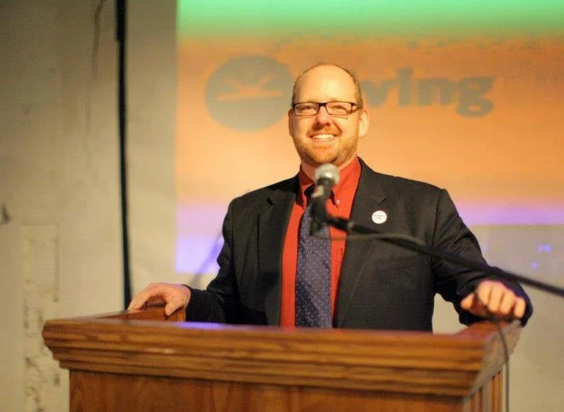 A picture of Jamie in glasses, a dark suit, and a red shirt standing behind a wooden podium, smiling at a microphone, with a colorful projection in the background.