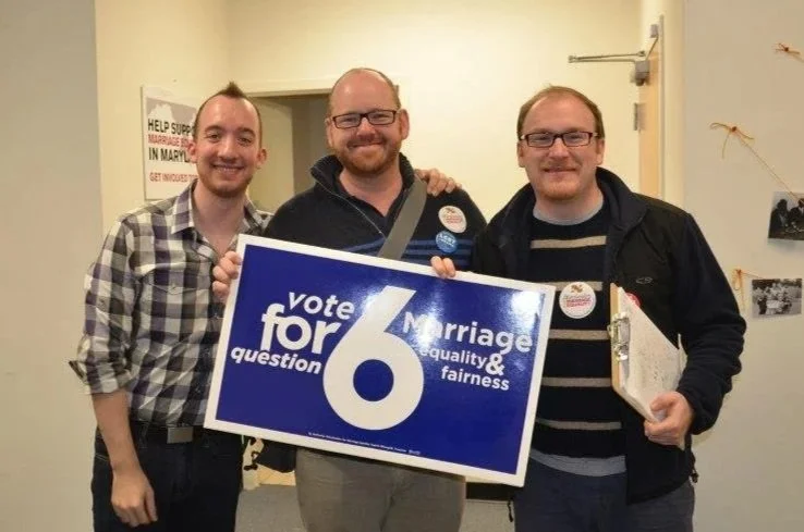 Jamie and Sean with their friend Tom at a marriage equality canvas; one holding a large sign that reads 'vote yes on question 6 for marriage equality & fairness,' and the man on the right holding a booklet. All are smiling.