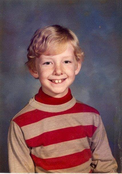 Jamie's kid photo with curly blonde hair and a missing front tooth, smiling, wearing a striped beige and red long sleeve shirt, against a blue gradient background.