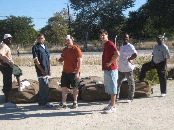 Group of six men standing outdoors near backpacks or duffel bags with trees and a fence in the background.