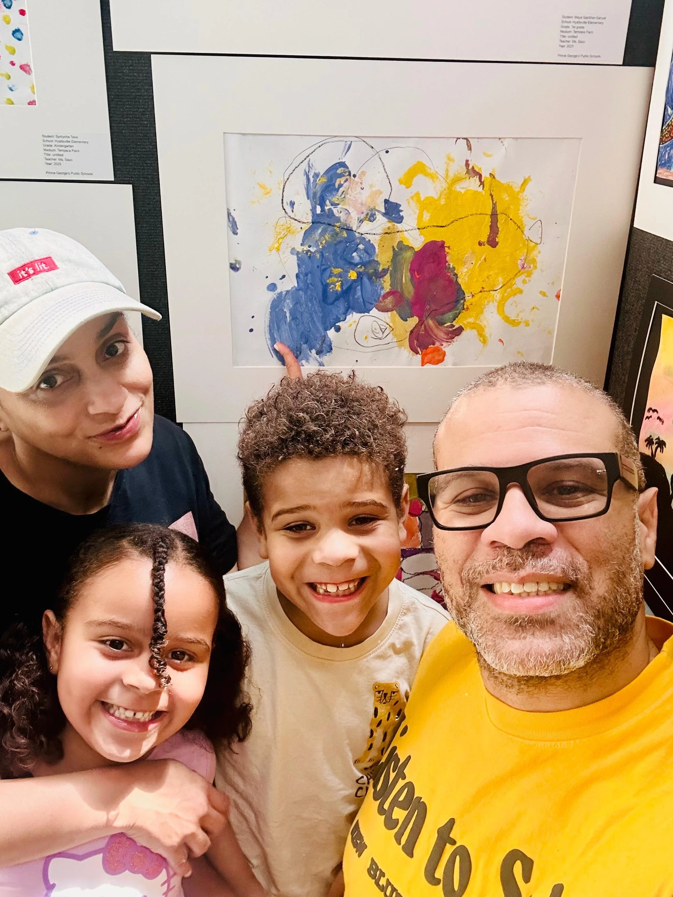 A group of five people, including two women and three children, smiling for a selfie in front of colorful artwork displayed on a wall. The artwork appears to be an abstract painting with blue, yellow, and red splashes.
