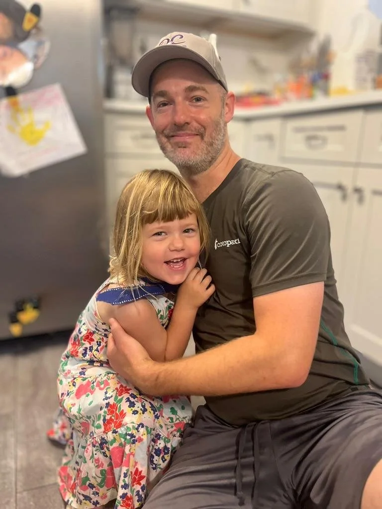 A man and young girl are sitting close together, smiling and hugging in a kitchen. The man is wearing a gray t-shirt and a baseball cap, and the girl is wearing a colorful, floral dress.