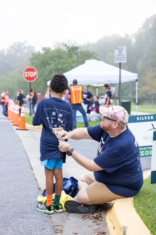 Jamie sitting on a curb and  handing a medal to a young boy at an outdoor event. In the background, there is a tent, a stop sign, and several people.