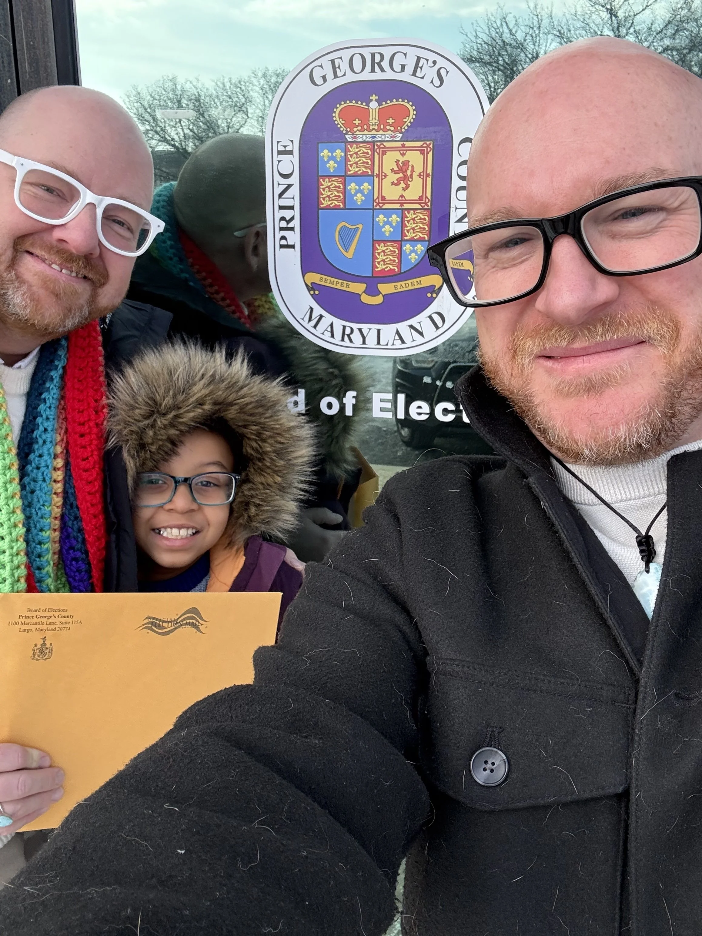Group of people taking a selfie in front of a Prince George's County, Maryland election board sign. One person is holding a yellow envelope, and a girl in a purple coat with a fur-lined hood is smiling in the center.