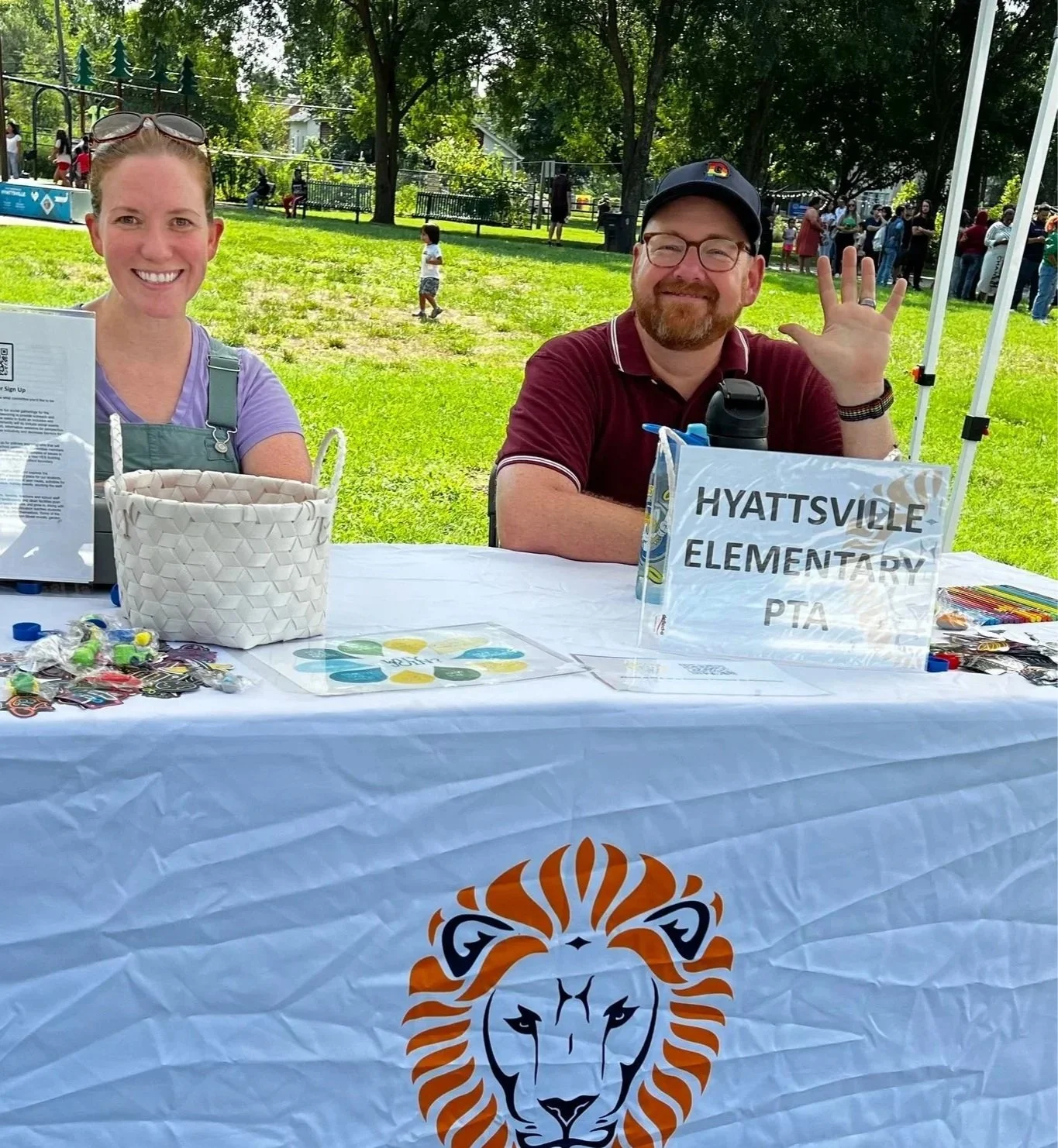 Two volunteers sitting at a table with a Lions Club International logo, outdoors at a community event. The woman on the left has sunglasses on her head and is smiling, the man on the right is waving and smiling, with a sign that reads 'HYATTSVILLE ELEMENTARY PTA' on the table.