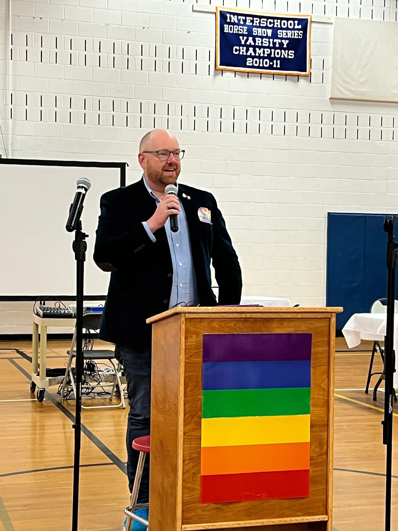 Jamie is accepting an award for Volunteer of the Year with Rainbow families. He's wearing a blue jacket and speaking at a podium with a rainbow flag in a school gym.