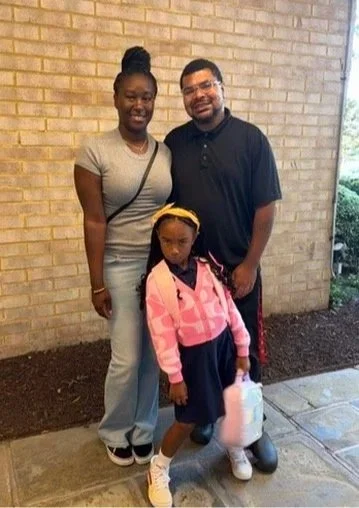 A family of three posing outside, with a woman, a man, and a young girl standing in front of a brick wall.