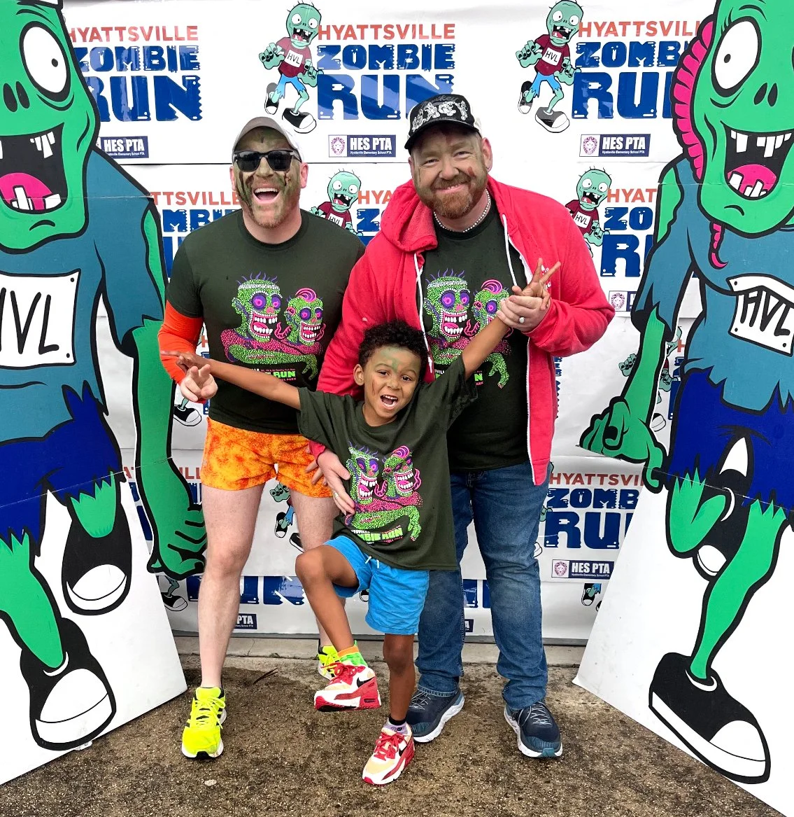 Jamie, Sean, and their son as zombies at the Hyattsville Zombie Run event, with cartoon zombie cutouts and a themed backdrop in the background.