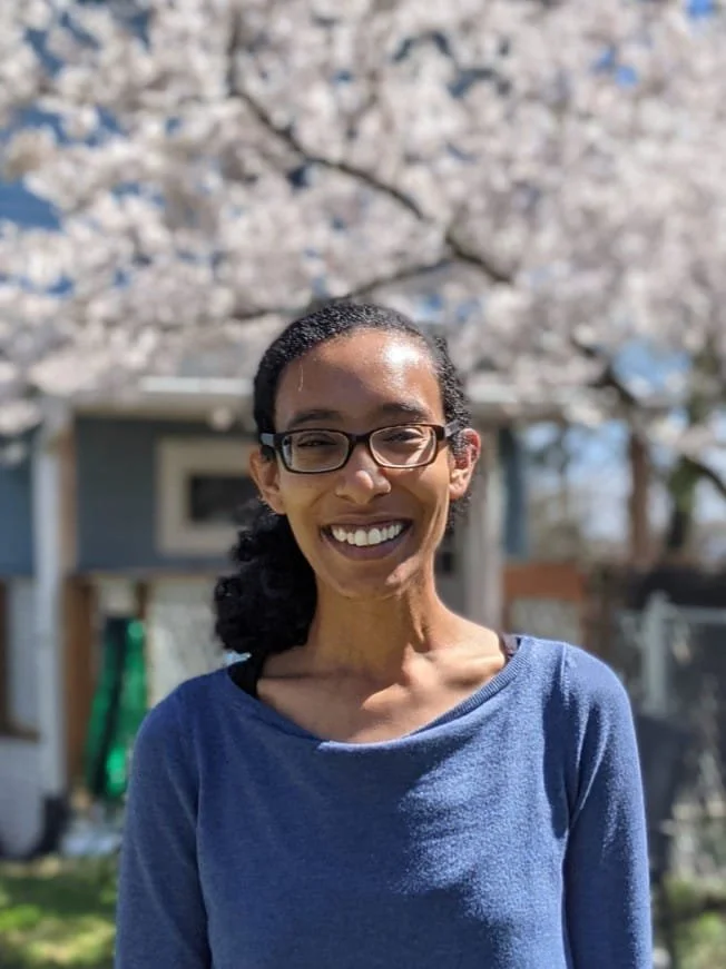 A woman with glasses and dark hair in a ponytail smiling outdoors with blossoming trees and a house in the background.