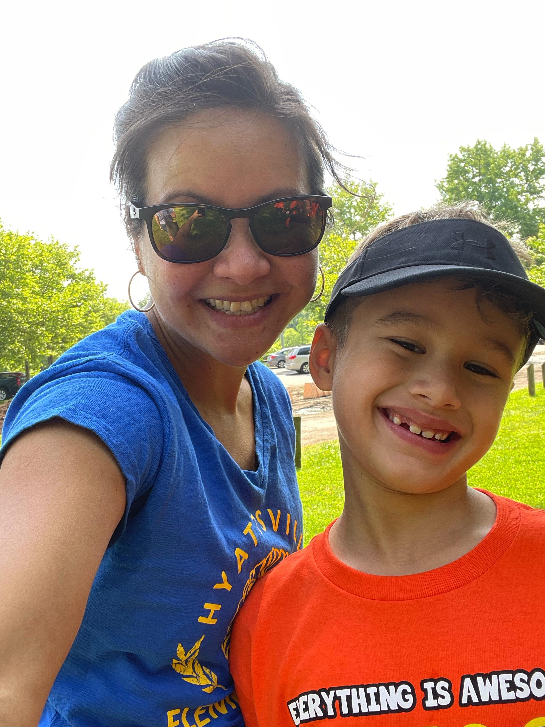 A woman and a young boy smiling outdoors on a sunny day. The woman is wearing sunglasses and a blue T-shirt, and the boy is wearing a black cap and an orange T-shirt. They appear to be happy and enjoying their time outside.