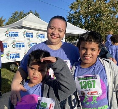 Three people smiling at a running event, with a woman in the middle and two children, all wearing race bibs and blue shirts, outdoors on a sunny day.