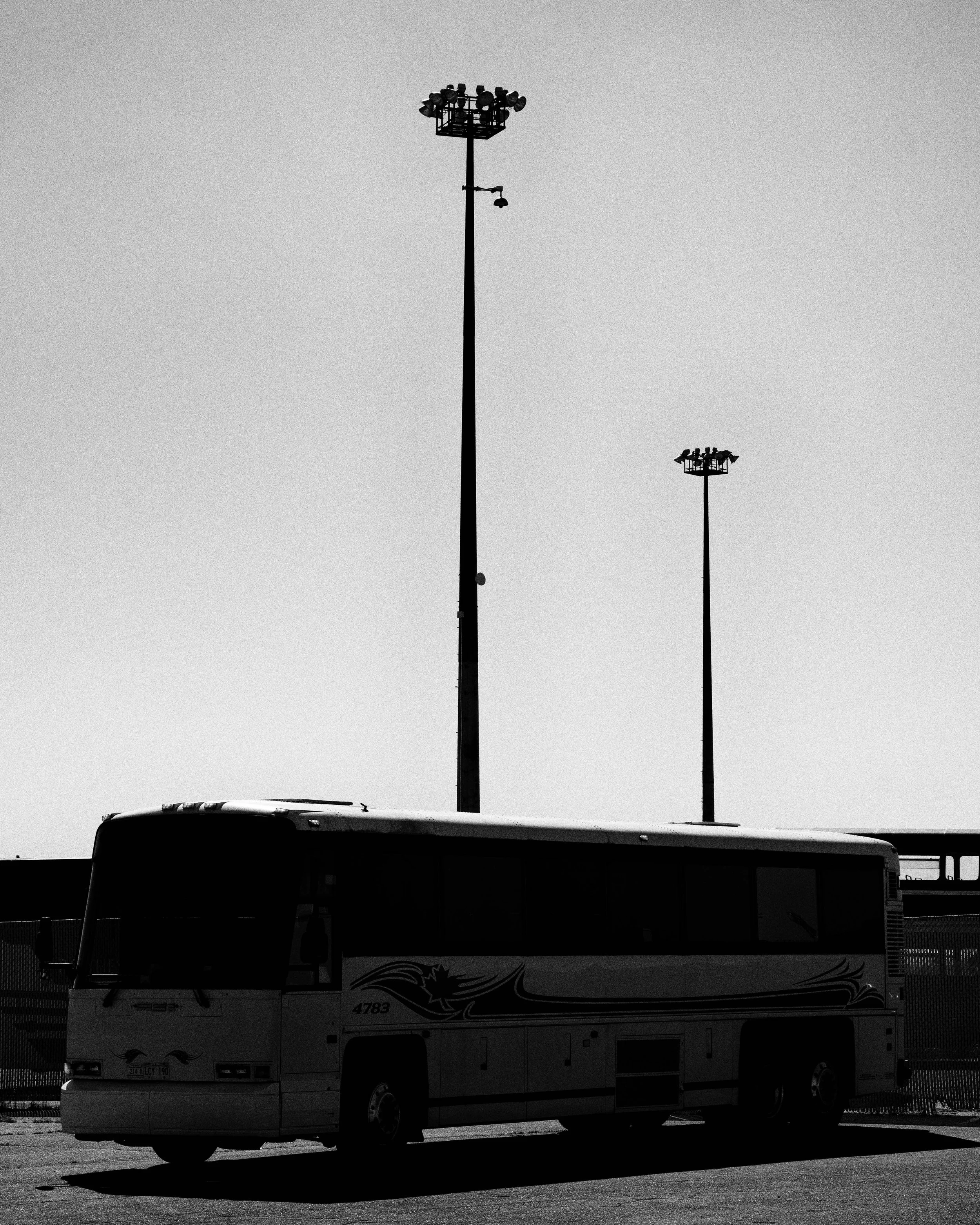 Black and white photo of a bus parked on a street with two tall stadium or parking lot light poles in the background.