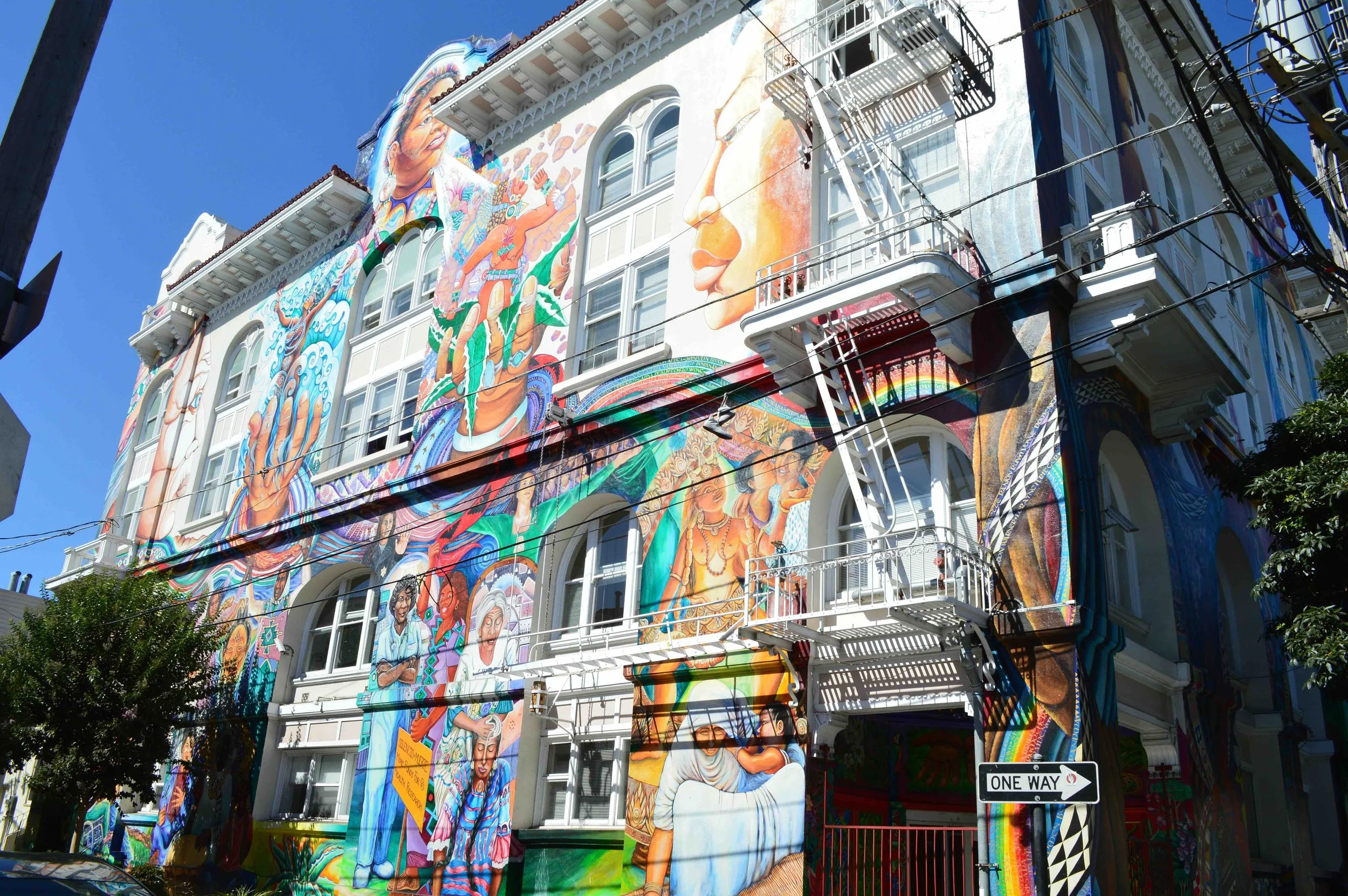Colorful mural of diverse people and cultural figures painted on the side of a multi-story building, with fire escape stairs and a street sign in foreground.